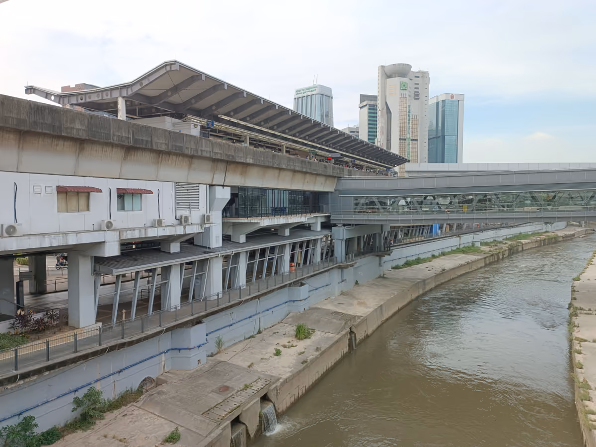 An elevated rail station with a long curved roof beside a concrete river channel, with a covered footbridge and office towers behind