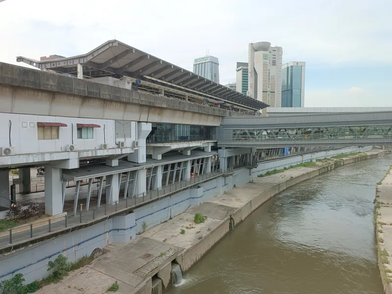An elevated rail station with a long curved roof beside a concrete river channel, with a covered footbridge and office towers behind