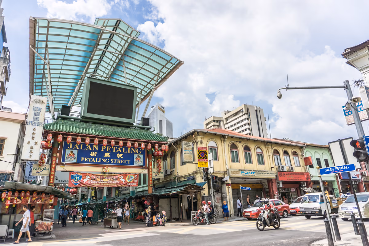 A street corner in Chinatown, with a green-tiled gateway sign reading Jalan Petaling and Petaling Street under a large glass canopy, red lanterns and two-storey shophouses
