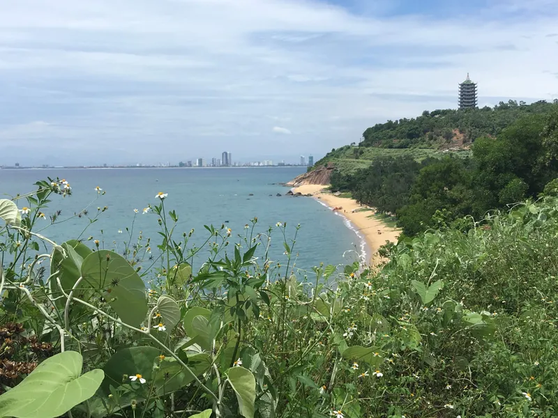 A green hillside above a small sandy cove, with the many-tiered tower of the Linh Ung Pagoda on the hill and the Da Nang skyline across the bay