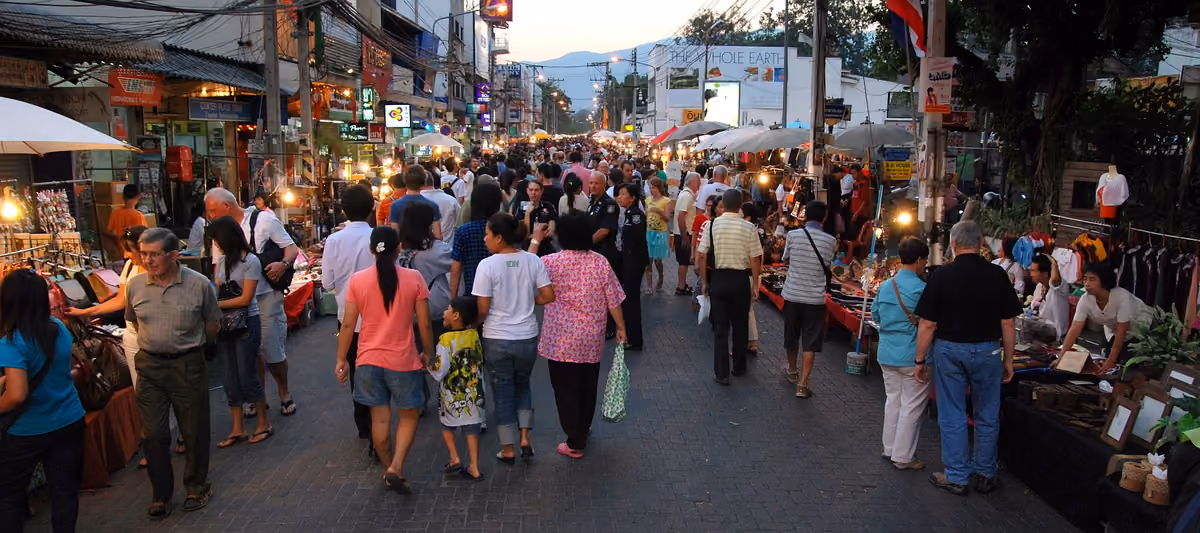 A wide street at dusk, crowded with people walking between rows of market stalls under umbrellas, with mountains on the horizon
