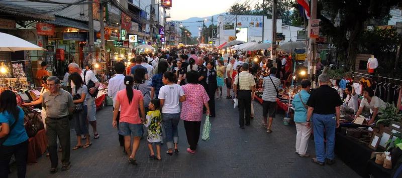 A wide street at dusk, crowded with people walking between rows of market stalls under umbrellas, with mountains on the horizon