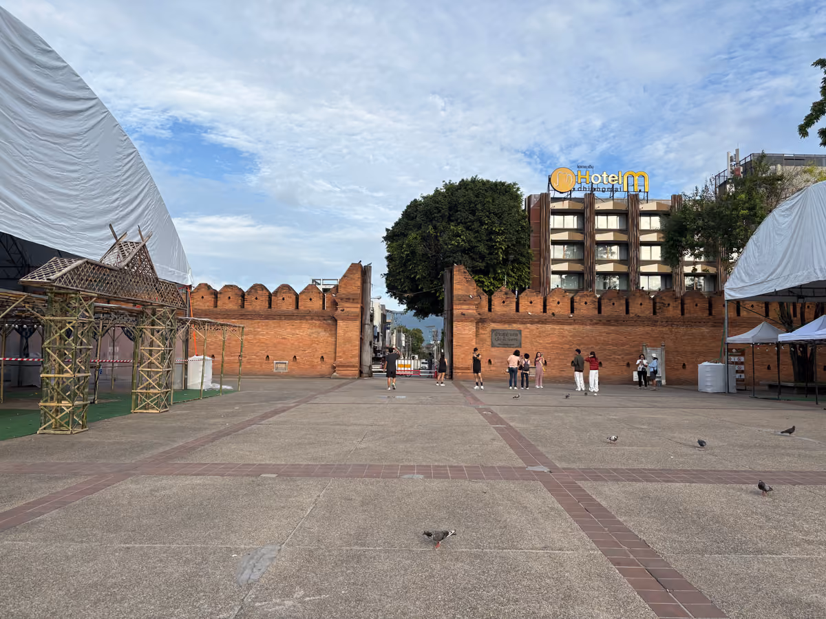 A wide paved square in front of a red brick city wall with battlements and an open gateway, with a few people walking in the square