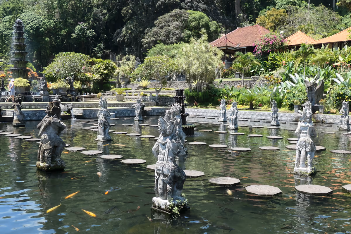 Carved stone guardian statues standing among round stepping stones in a pond with orange koi, with gardens, a tiered fountain and red-roofed pavilions behind