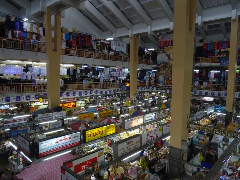 Several floors of a covered market seen from an upper gallery, with clothes hanging from the balconies and rows of stalls with Thai signs on the floor below