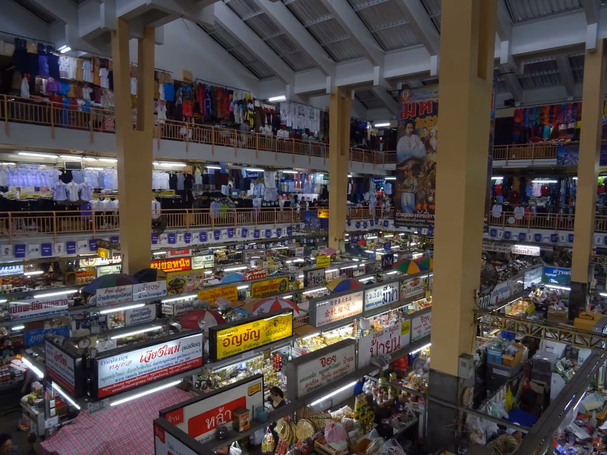 Several floors of a covered market seen from an upper gallery, with clothes hanging from the balconies and rows of stalls with Thai signs on the floor below