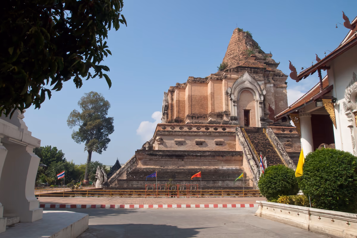 A huge ruined brick chedi with a naga staircase up one face, small coloured flags at its base and a white temple building on the right