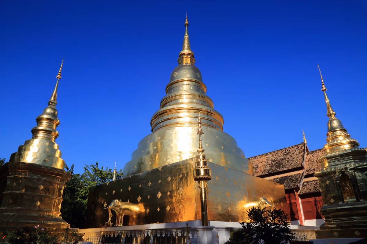 A large gold-covered chedi with a tall spire under a deep blue sky, two smaller gold chedis on either side and a gold elephant statue at its base