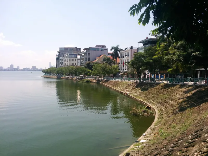 A curving stone embankment along West Lake, with trees and four- and five-storey houses along the shore and tall buildings in the hazy distance
