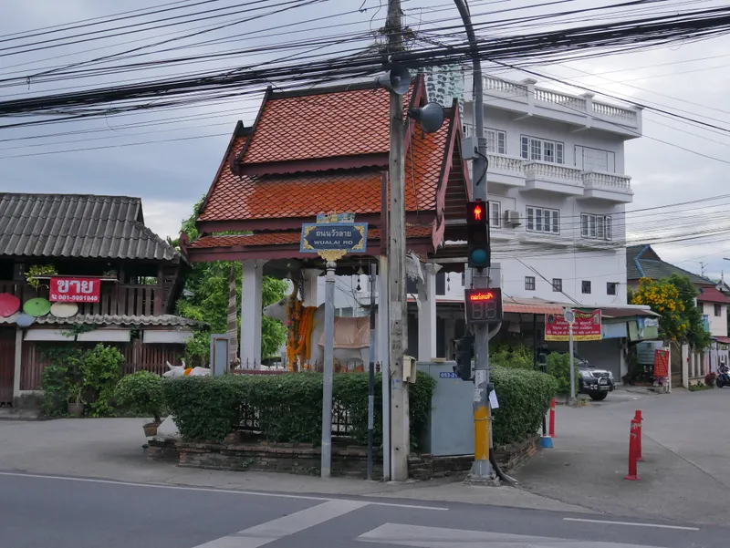A street corner with a red-tiled open pavilion behind a low hedge, a blue street sign reading Wualai Rd in Thai and English, traffic lights and white shophouses behind